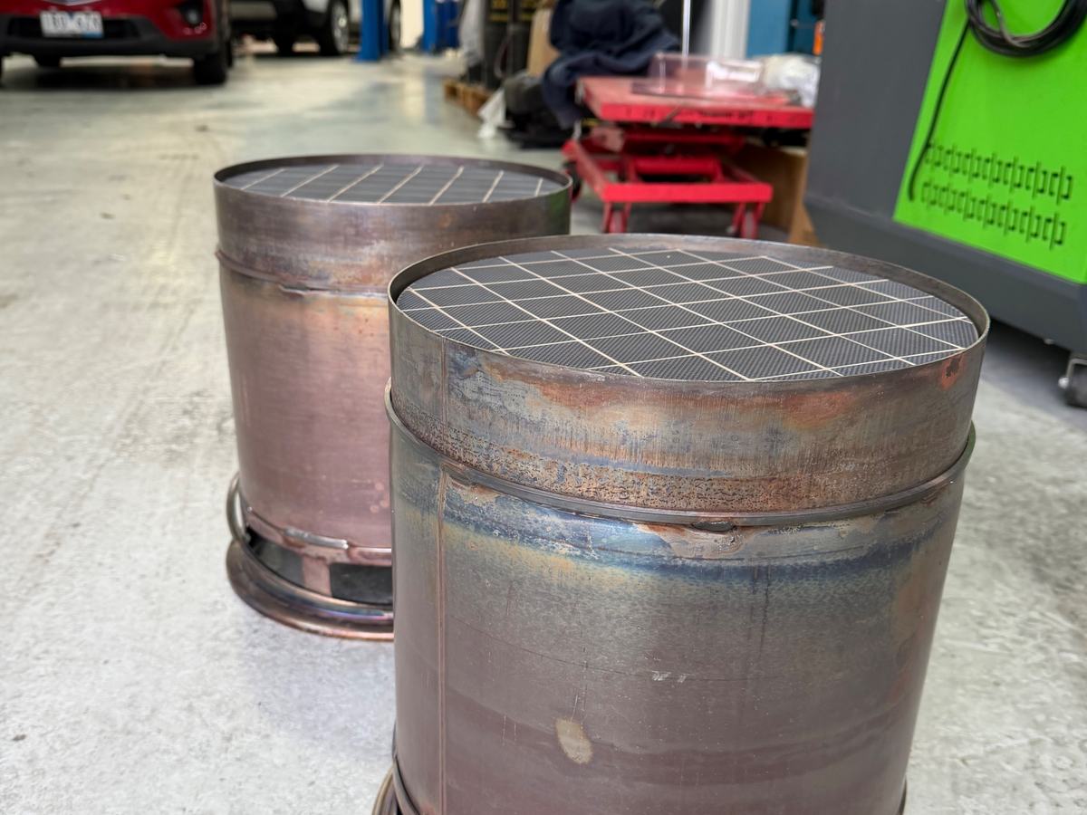 Two large truck diesel particulate filter canisters standing on the workshop floor with cleaned substrate faces visible, the cleaning cabinet beside them.